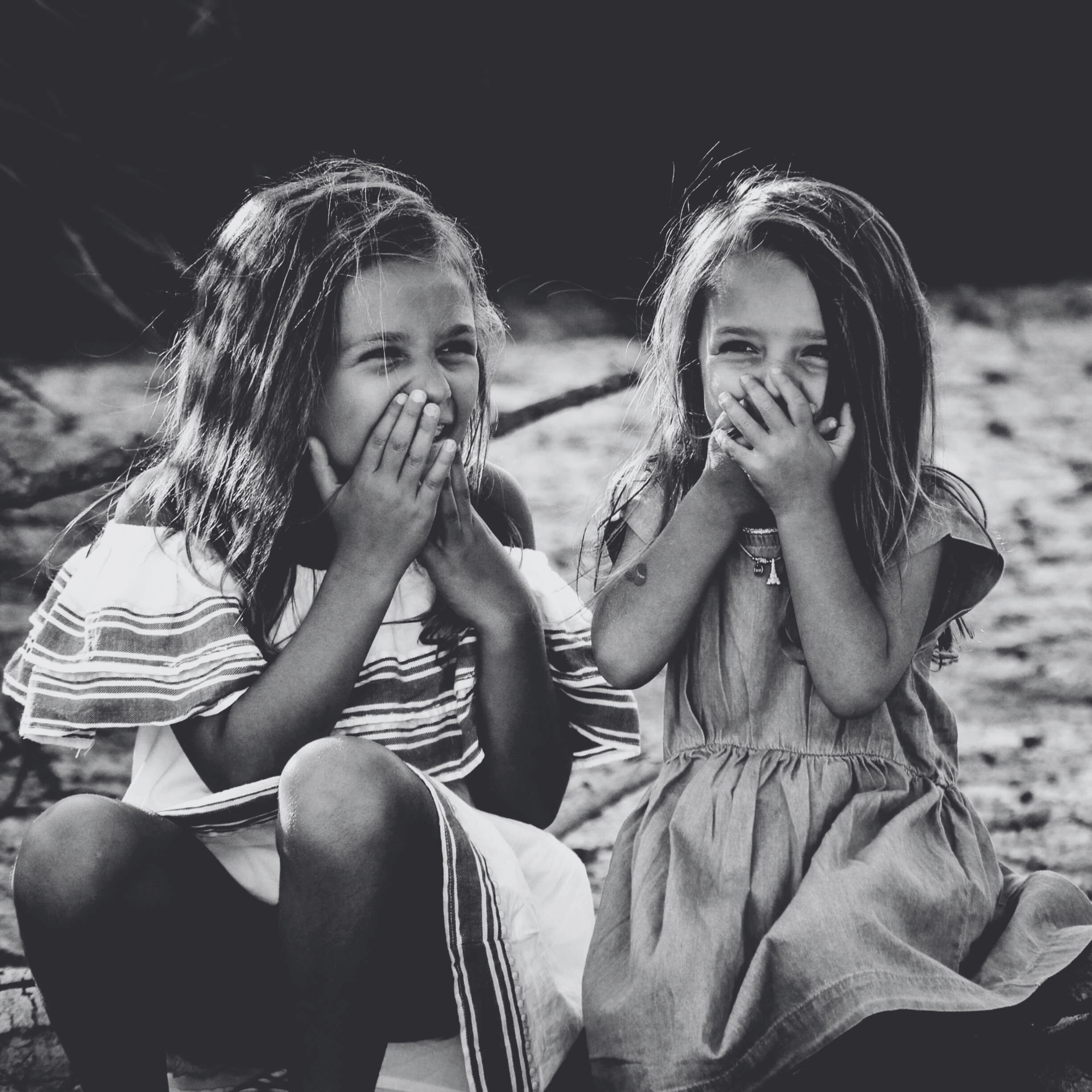 Two young girls sitting outdoors with their hands over their mouths.