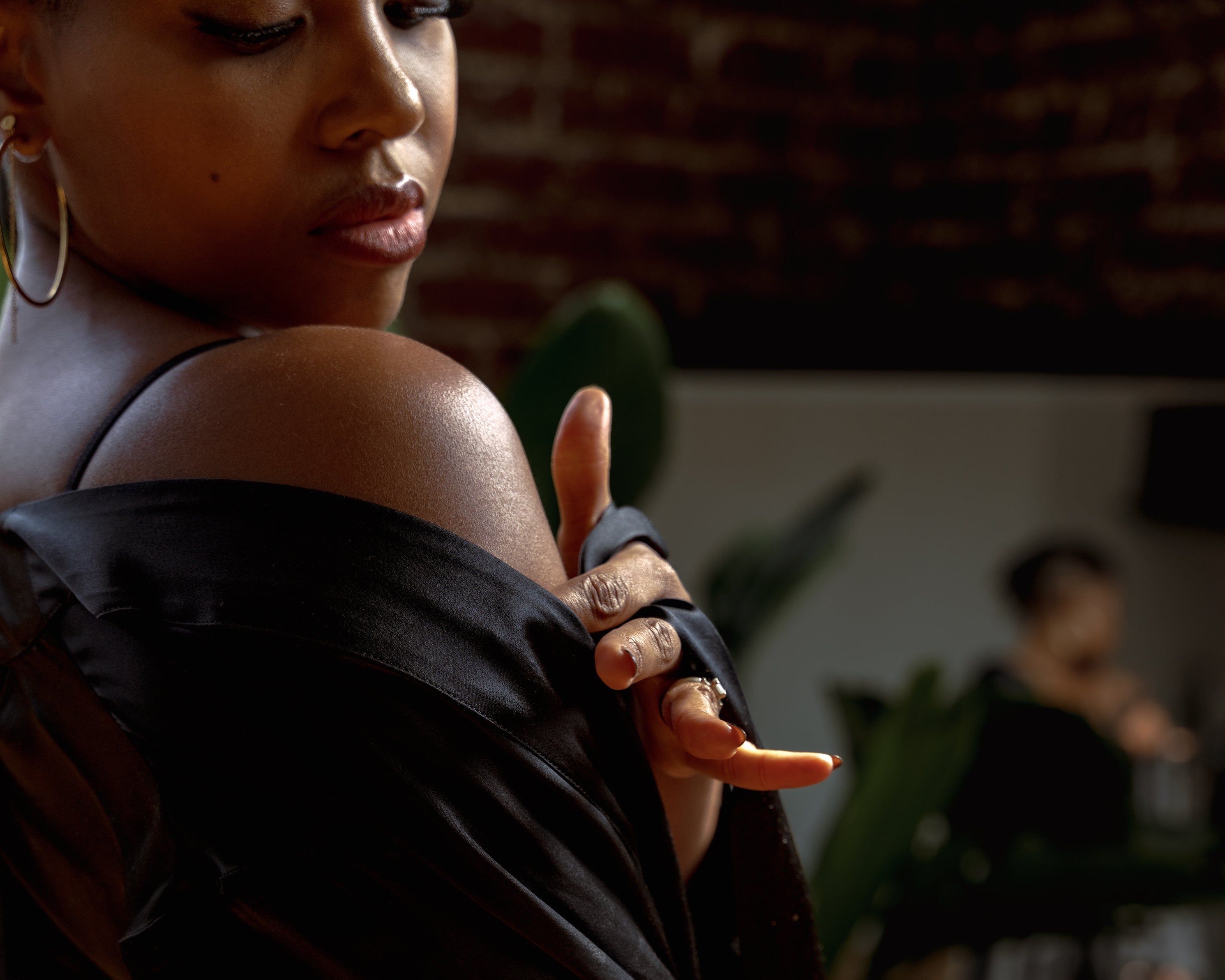 Woman wearing a black off-shoulder top in a dimly lit room with plants.