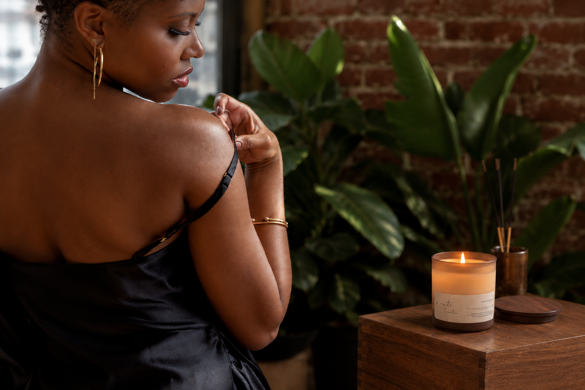 A woman in a black satin top is shown from behind in a warmly lit indoor setting with a brick wall and large green plants. In the foreground, a lit candle in a clear glass jar sits on a wooden surface next to a reed diffuser.