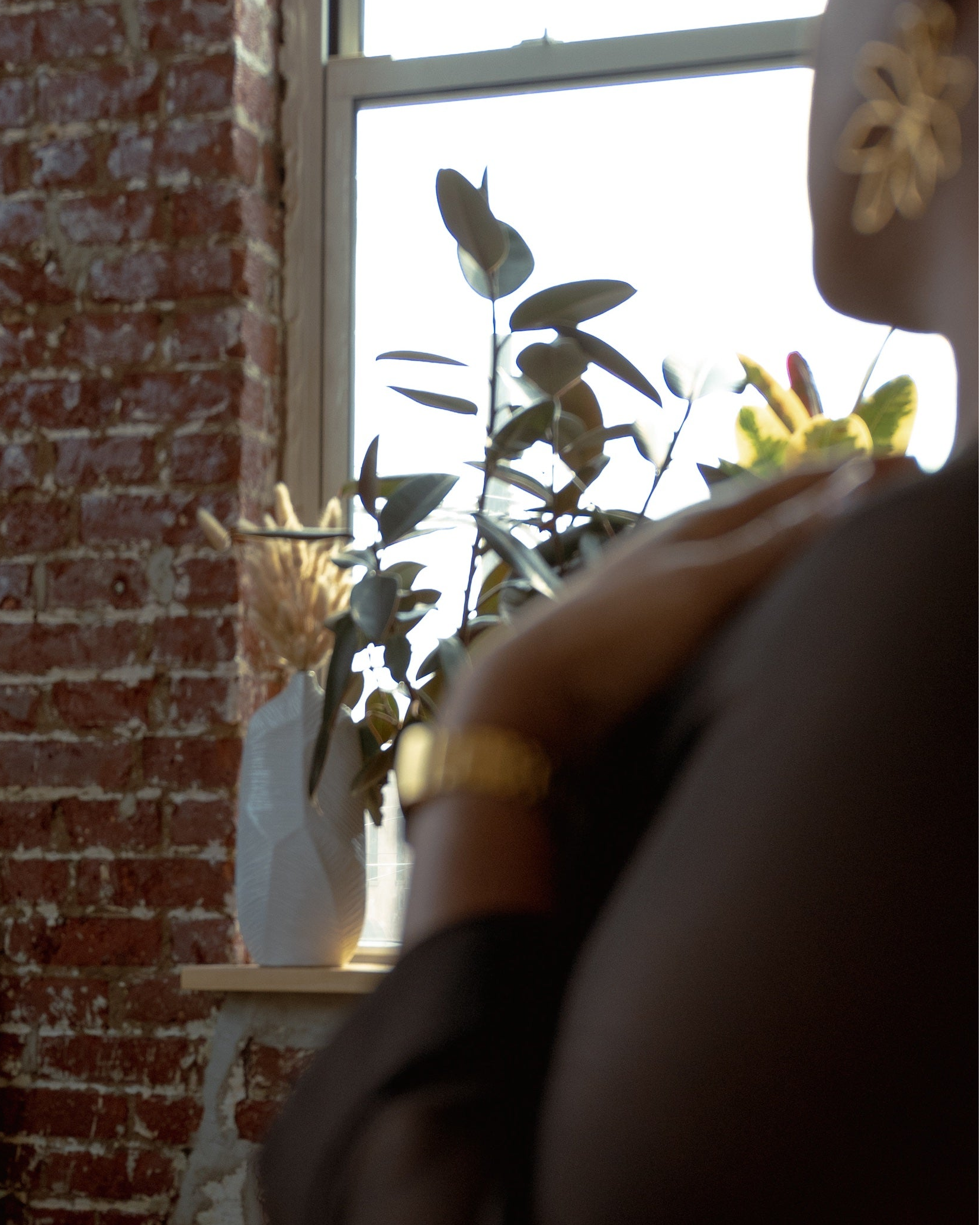 Person with a blurred face standing in a room with brick wall and plants wearing je mérite