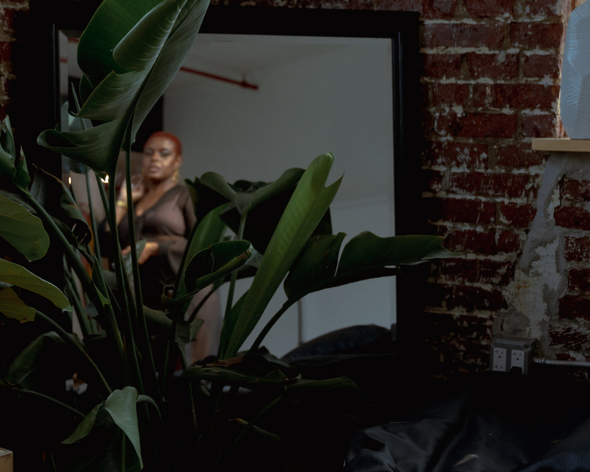 Person standing in a room with a large mirror and plant in the foreground.