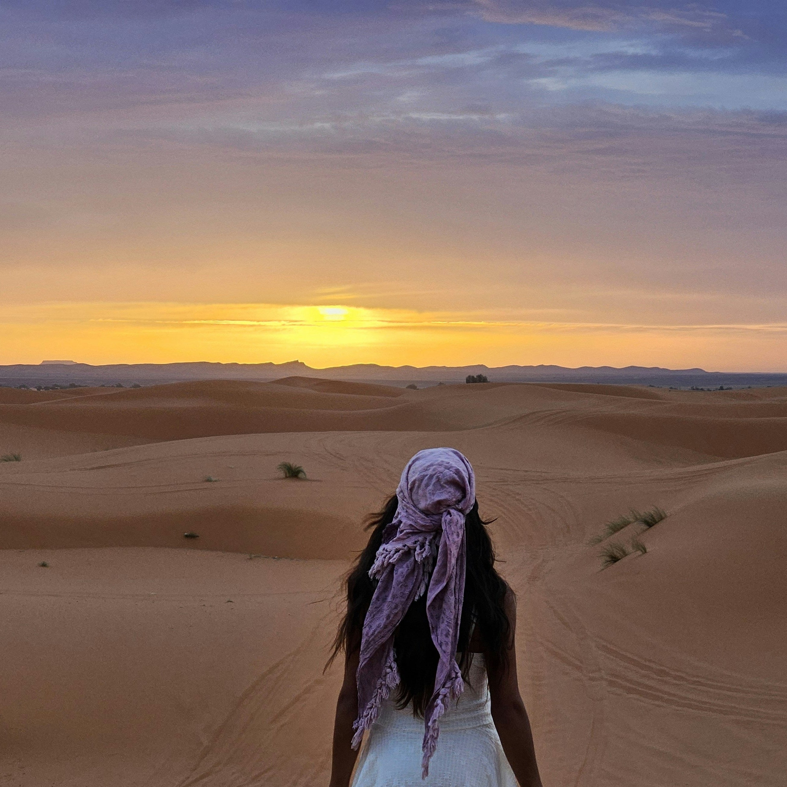 A woman walking barefoot across desert sand dunes at sunset, seen from behind, wearing a white dress and a purple patterned headscarf.