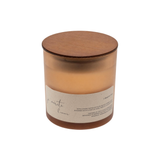 A soy wax candle in a translucent amber glass jar with a round wooden lid and a cream paper label, shown on a plain white background.