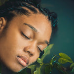 Person with braided hair resting their head on green leaves against a dark background