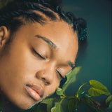 Person with braided hair resting their head on green leaves against a dark background