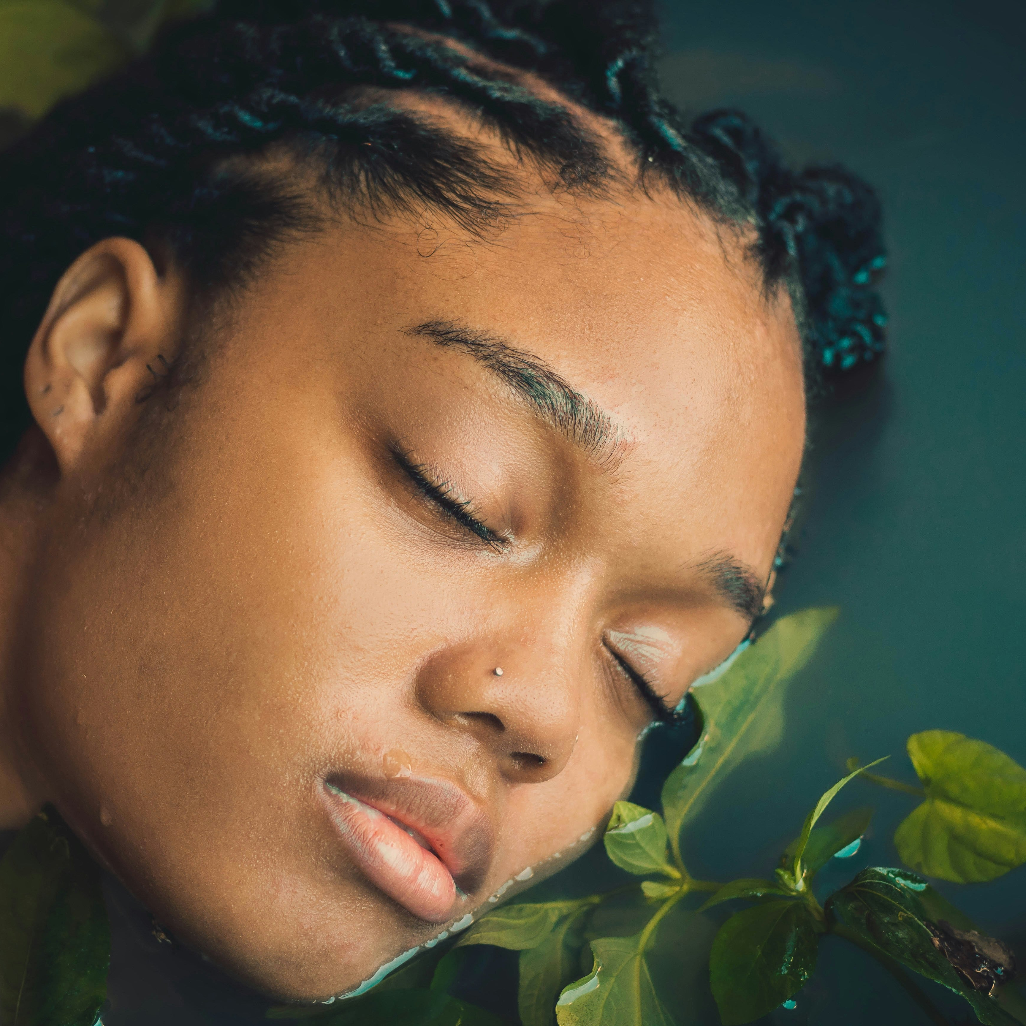 Person with braided hair resting their head on green leaves against a dark background