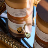 Two cylindrical je merite candle vessels with labels on a reflective surface, with a gold necklace.
