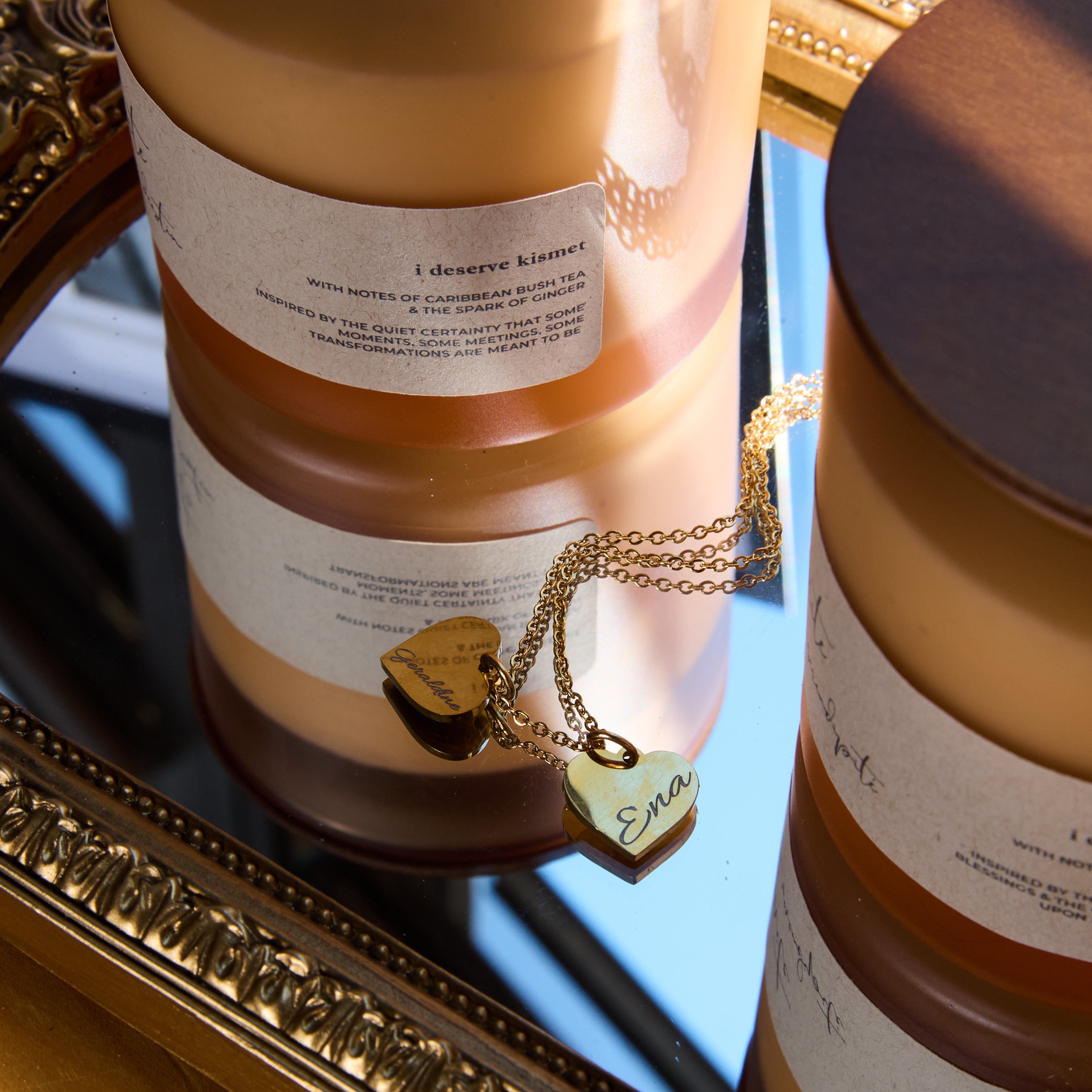 Two cylindrical je merite candle vessels with labels on a reflective surface, with a gold necklace.