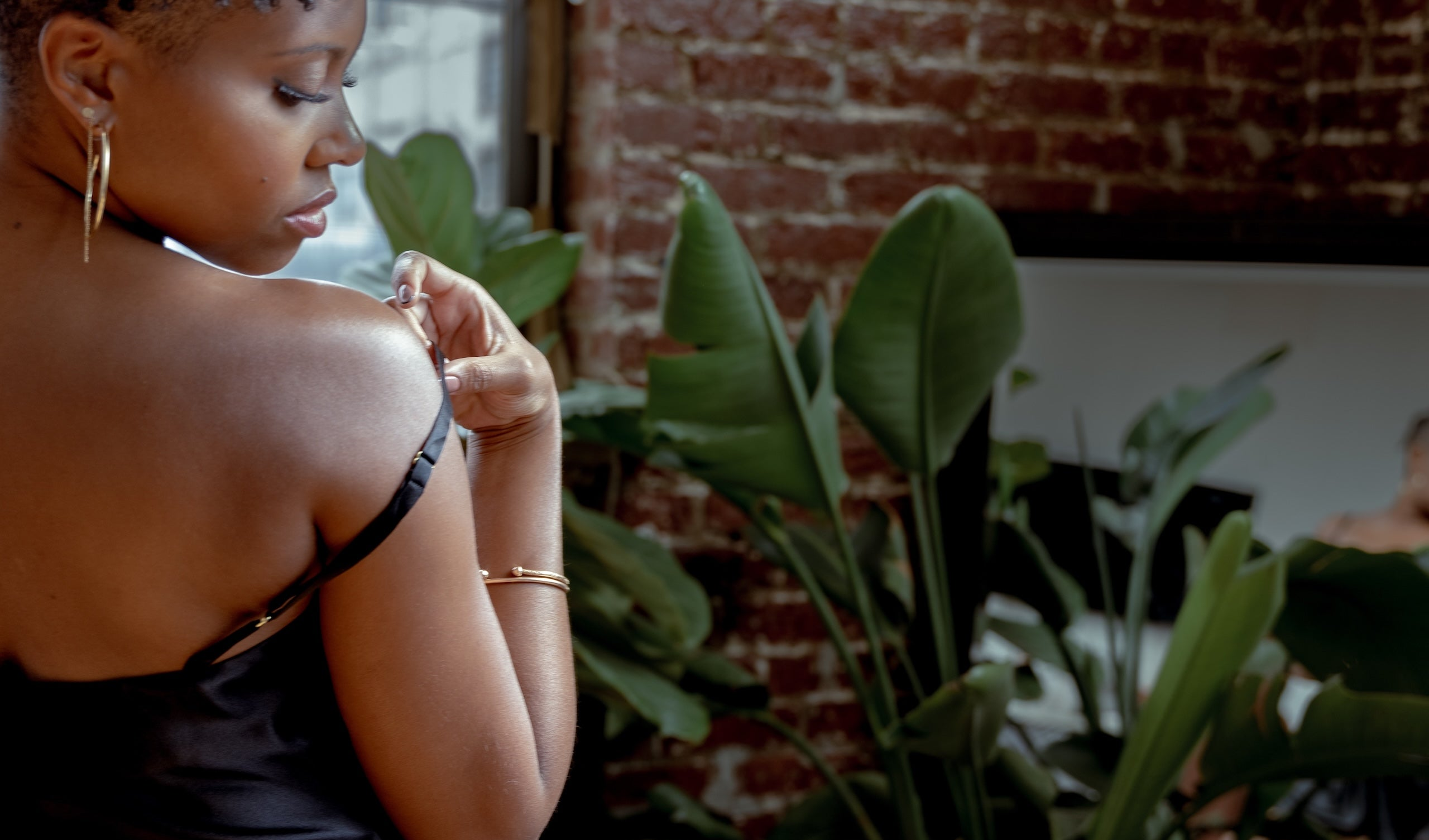 A woman in a black satin slip dress with thin straps, shown from behind and side as she adjusts the strap on her shoulder. The setting is a stylish room with a brick wall, large green houseplants, and a mirror reflecting another person.