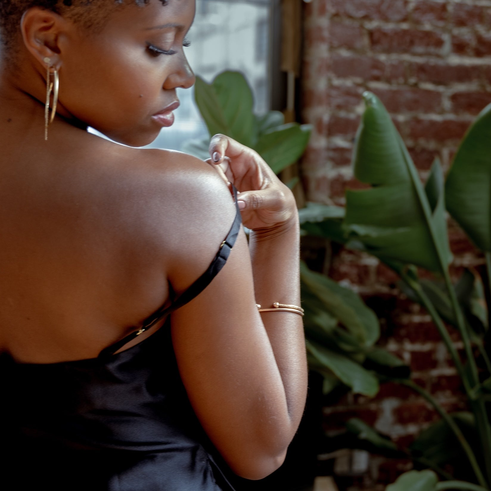 A woman in a black satin slip dress with thin straps, shown from behind and side as she adjusts the strap on her shoulder. The setting is a stylish room with a brick wall, large green houseplants, and a mirror reflecting another person.