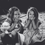 Two young girls sitting outdoors with their hands over their mouths.