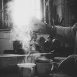Person pouring hot liquid from a pot into cups with steam rising, in a kitchen setting.